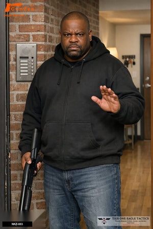 Man in black hoodie holding shotgun low ready, hand raised, in apartment foyer.